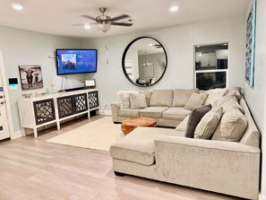 A living room with a beige sofa, a coffee table, a TV on a stand, a ceiling fan, and a large mirror. - Bertram Tranquility- 3 Bedroom 2 Bath Hideaway (Bertram)