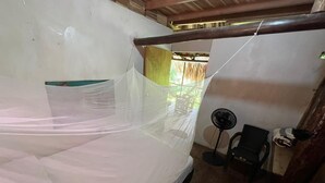 Une pièce avec un lit recouvert d’un drap blanc, un ventilateur sur pied et une chaise.