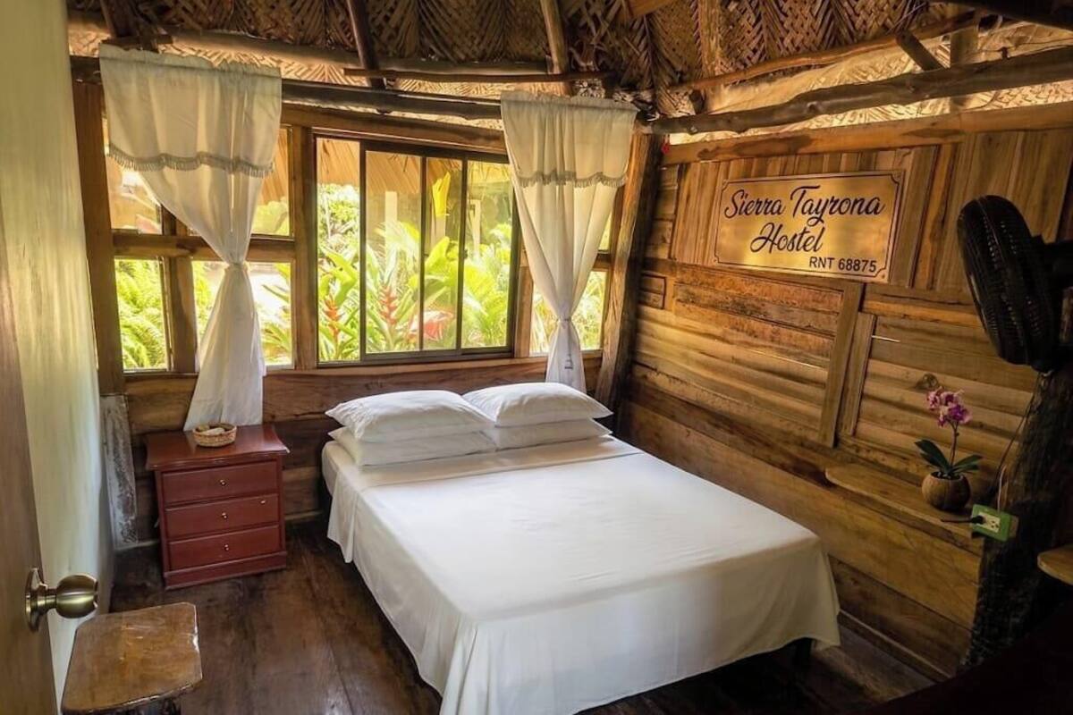 A wooden room with a bed, a nightstand, a window with curtains, and a sign for Sierra Tayrona Hostel.
