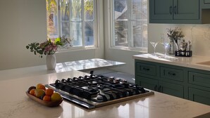 Een keuken met een gasfornuis, een fruitschaal, een bloemenvaas en een raam met jaloezieën.