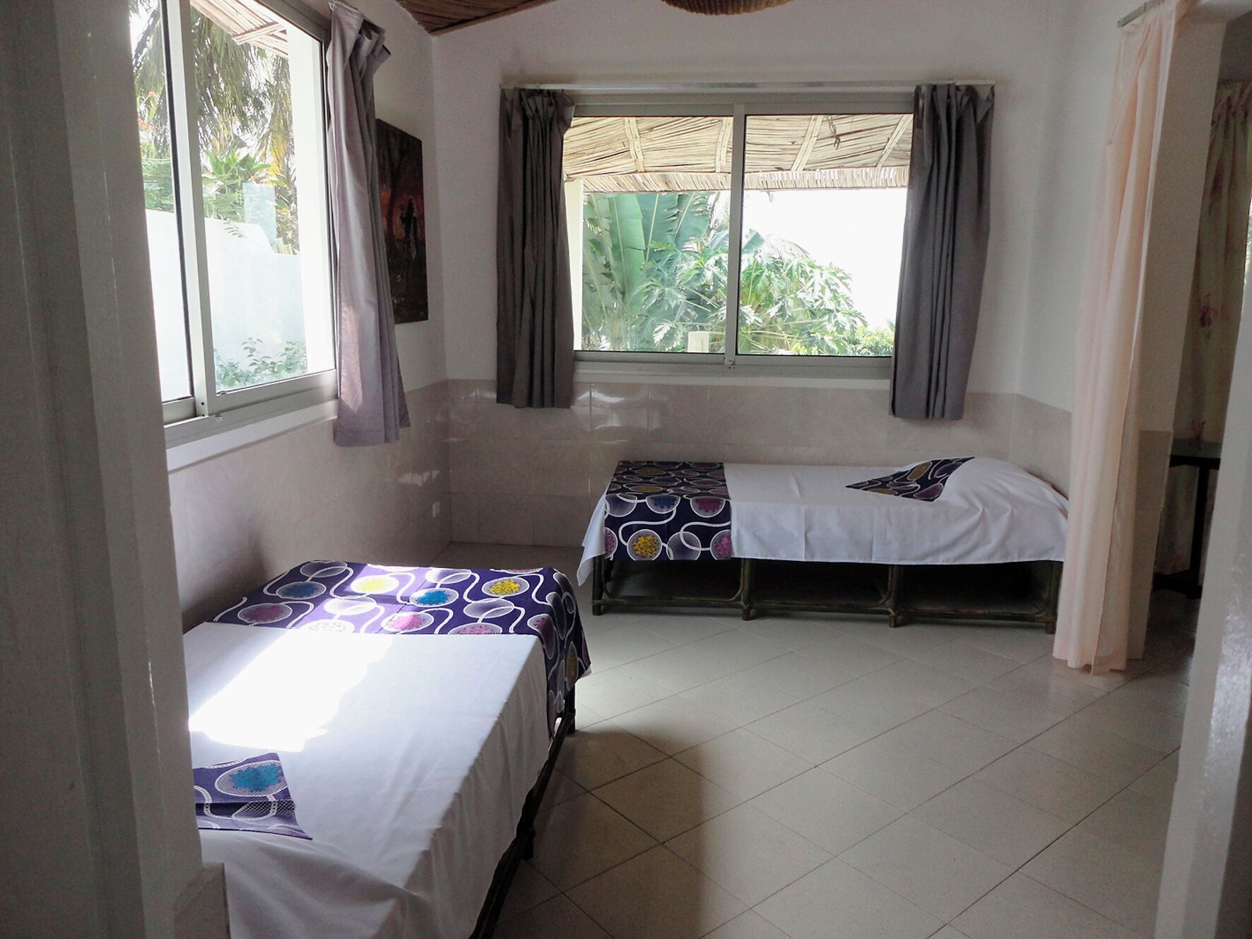 A room with two beds, a window with curtains, and a view of greenery outside.