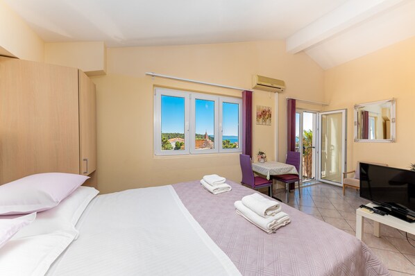 A hotel room with a bed, a desk with a TV, a chair, a table, and a sliding glass door leading to an outdoor area.