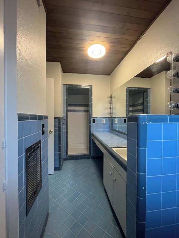 A narrow bathroom with blue tiles, a shower, and a mirror. - Sleepy Hollow Villa (Rosenberg)