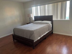 A bedroom with a wooden bed, a nightstand, a lamp, and a window with blinds. - Sleepy Hollow Villa (Rosenberg)
