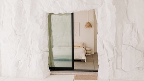 Une pièce avec un mur blanc à texture, un lit avec une literie blanche, un luminaire suspendu et une porte-fenêtre coulissante donnant sur une autre pièce.