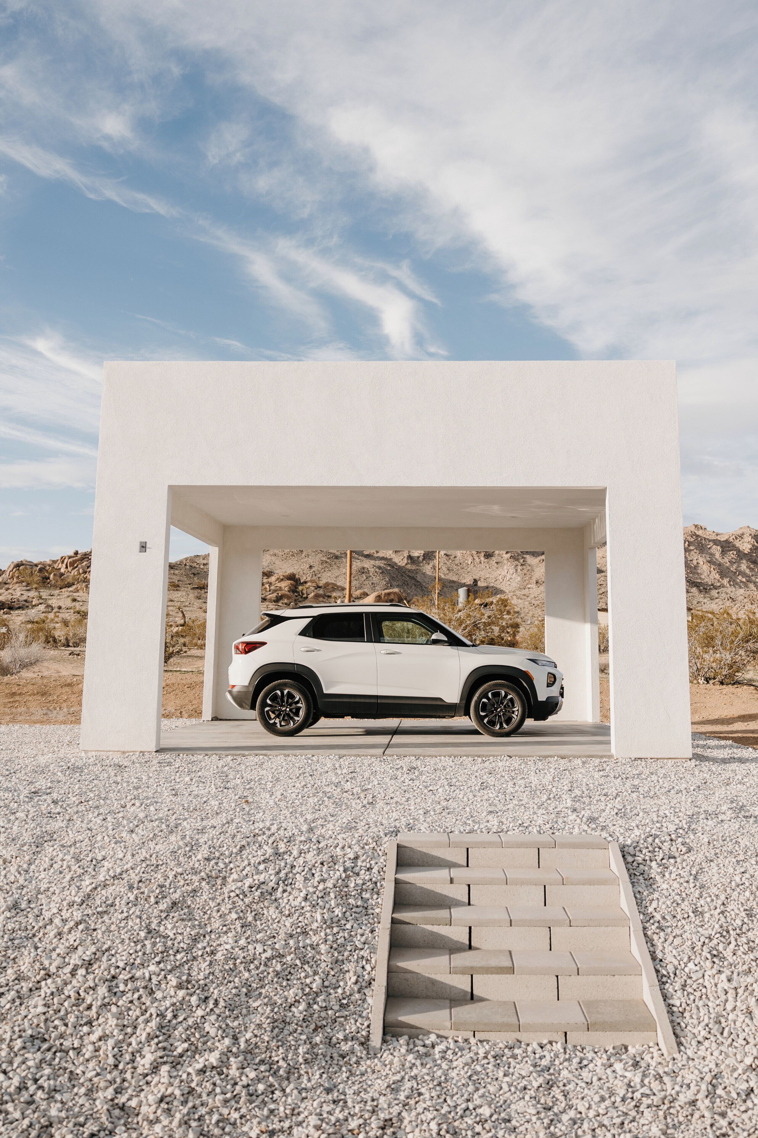 A white SUV parked under a white structure with a gravel ground and a mountainous background.