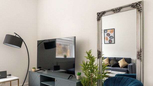 A modern living room with a flat-screen TV, a large ornate mirror, a blue velvet armchair, a small table, and a floor lamp.
