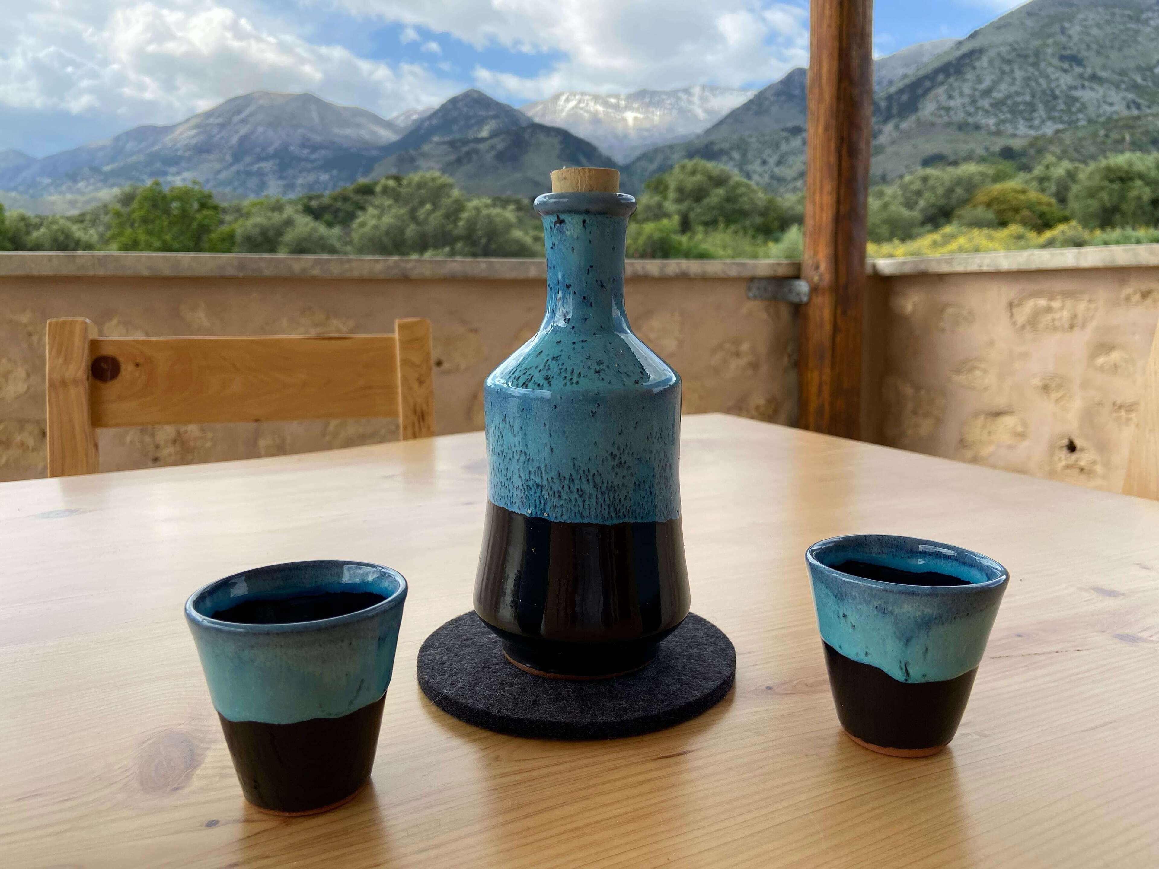 Een tafel met een fles en twee kopjes, met uitzicht op een bergachtig landschap.