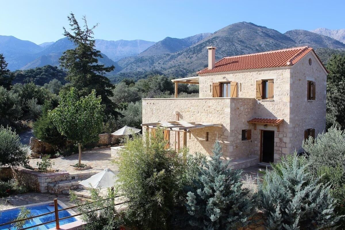 A stone house with a red-tiled roof, a pool, and a mountainous backdrop.