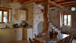 A rustic kitchen with stone walls, wooden beams, and a dining table with chairs. - Villa Ariadne - Stylish vacation home with all amenities (Melidoni)