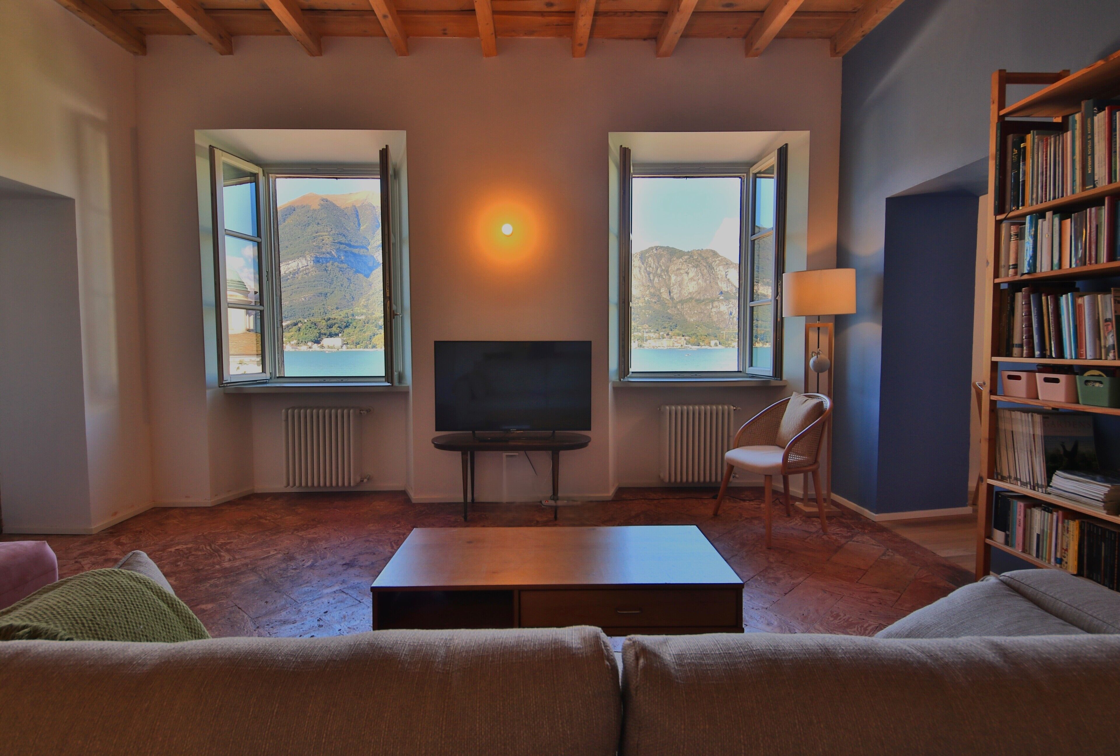 Una sala de estar con televisión, dos ventanas con vistas a la montaña, una estantería y una lámpara.