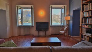 En stue med fjernsyn, to vinduer der ud til et bjerglandskab, en bogreol og en lampe.