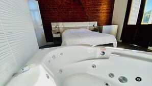 Une chambre d’hôtel moderne avec une grande baignoire blanche, un lit avec une literie blanche et un mur en briques en arrière-plan.