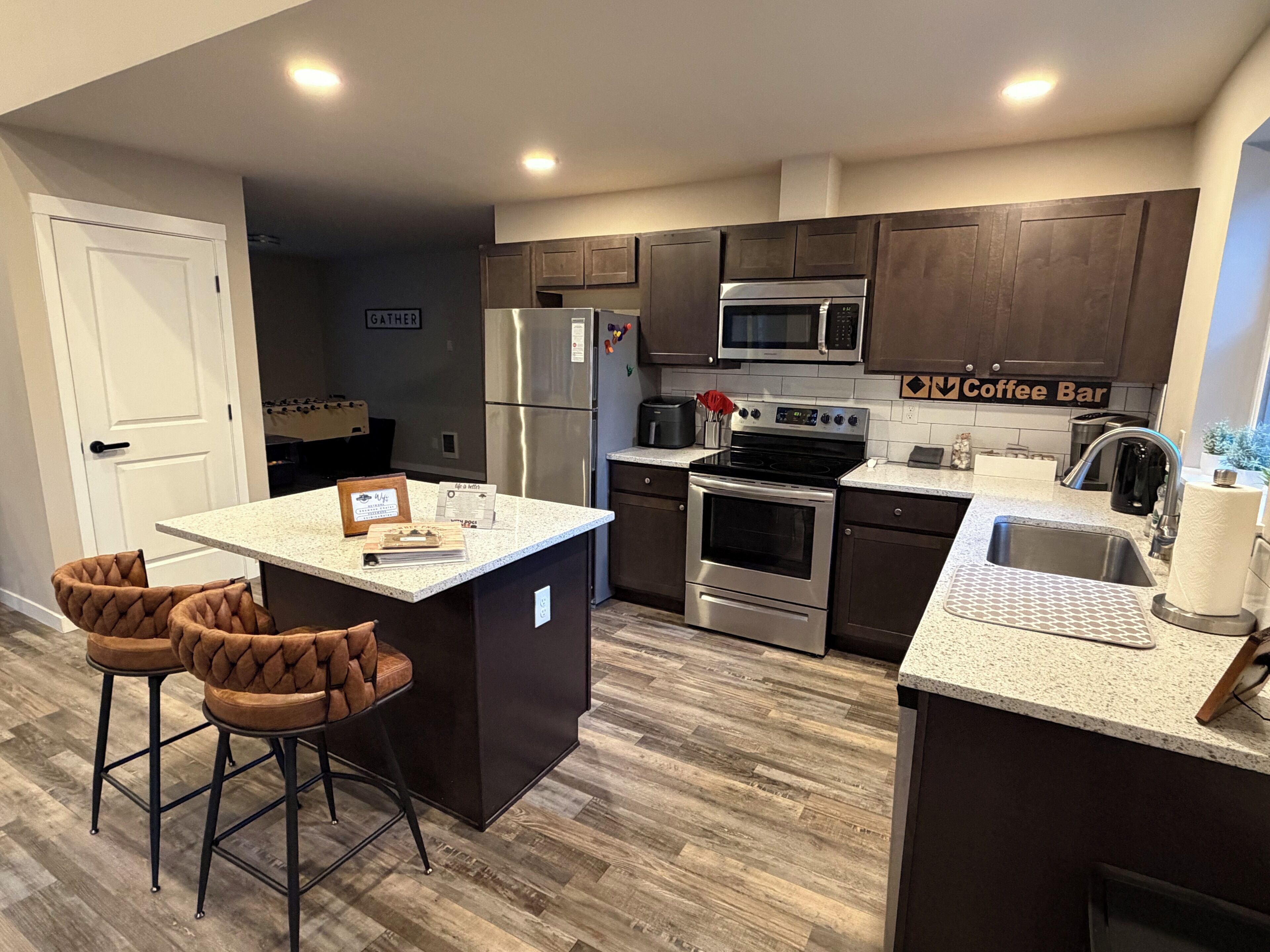 A modern kitchen with a central island, bar stools, a coffee bar sign, stainless steel appliances, and a dark wood floor.