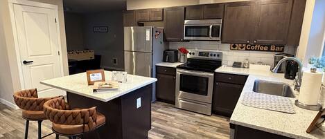 A modern kitchen with a central island, bar stools, a coffee bar sign, stainless steel appliances, and a dark wood floor.
