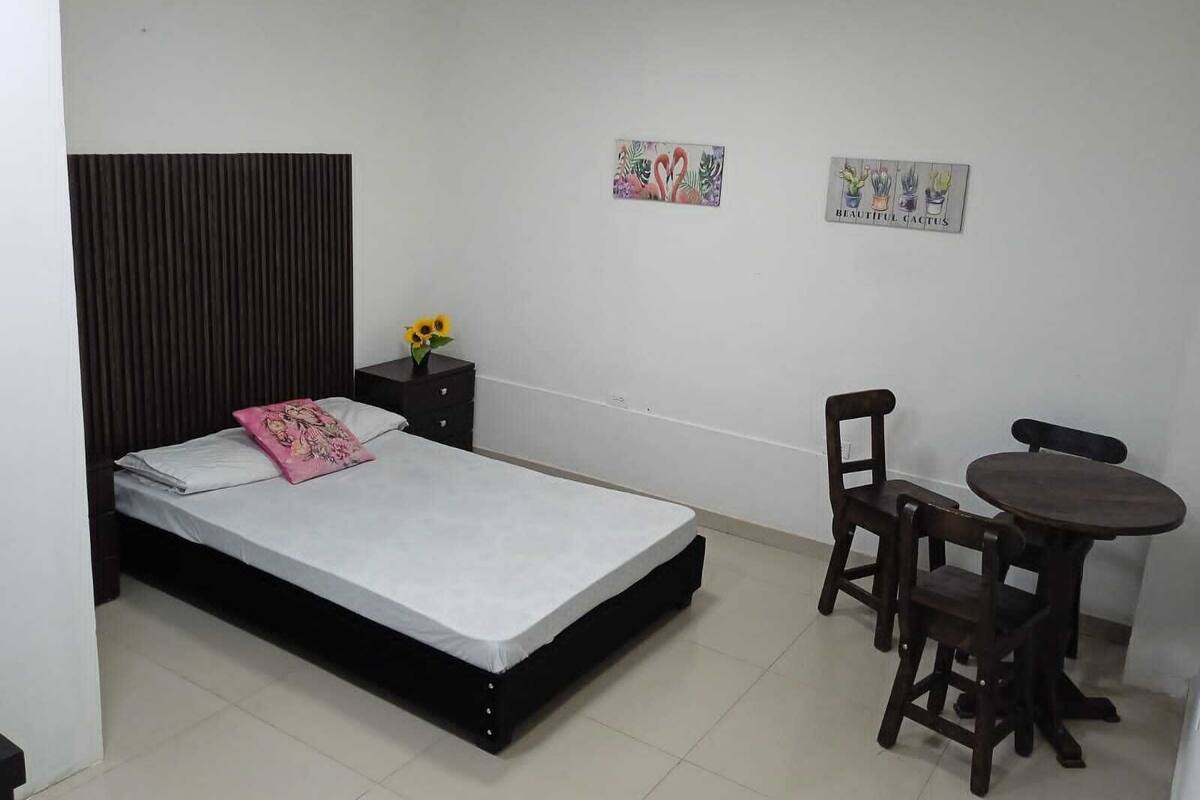 A bedroom with a bed, a nightstand, a chair, a small table, and two wall art pieces.