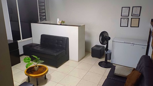 Ett kompakt vardagsrum med en svart soffa, ett litet träbord med en växt, ett vitt kylskåp och en golvfläkt.