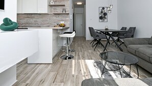 Una cocina moderna con armarios blancos, un revestimiento de pared de madera y una zona de comedor con mesa de vidrio y sillas tapizadas en gris.