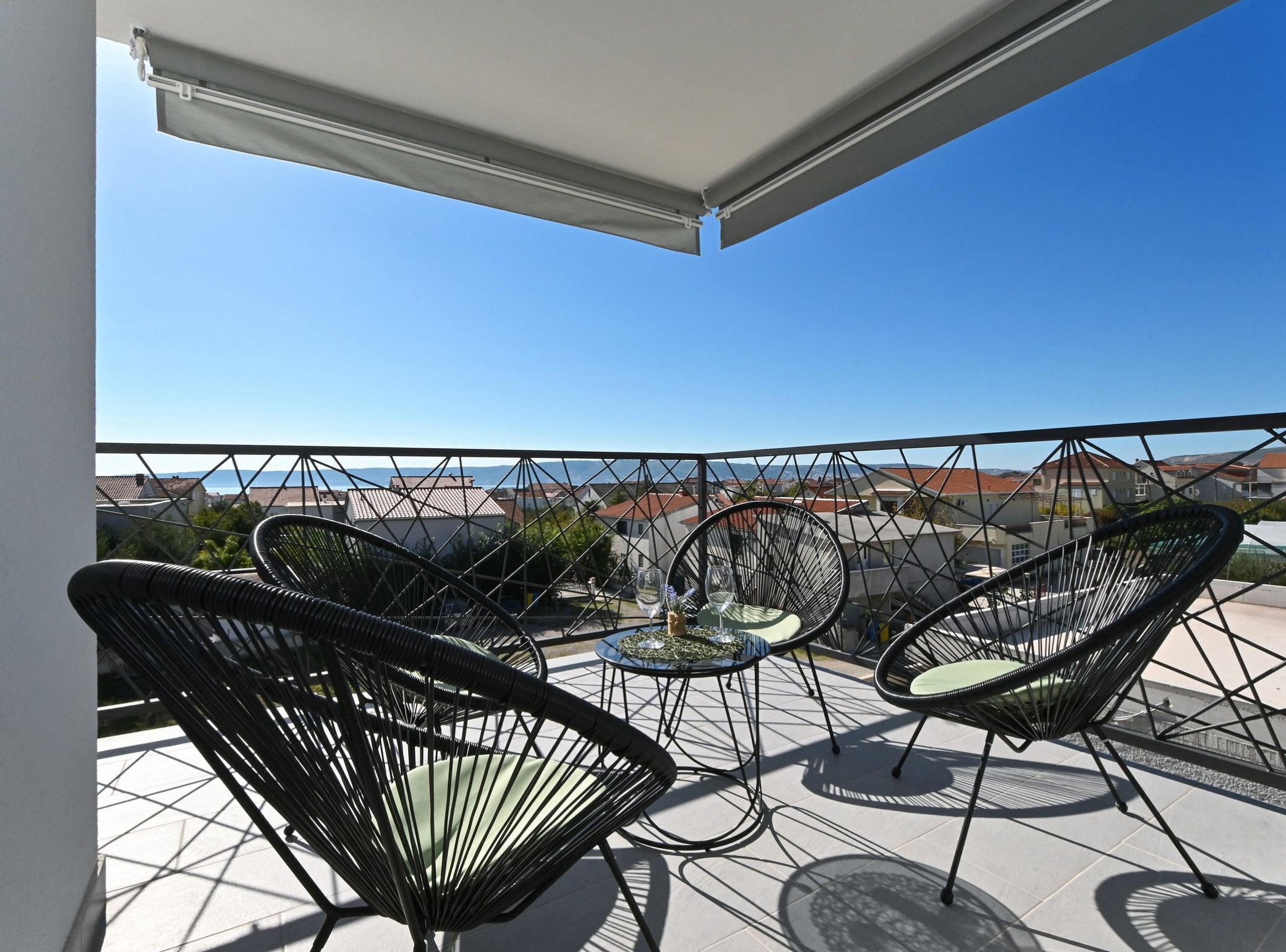 A balcony with a view of the sea, featuring black wireframe chairs and a small table with a glass top.