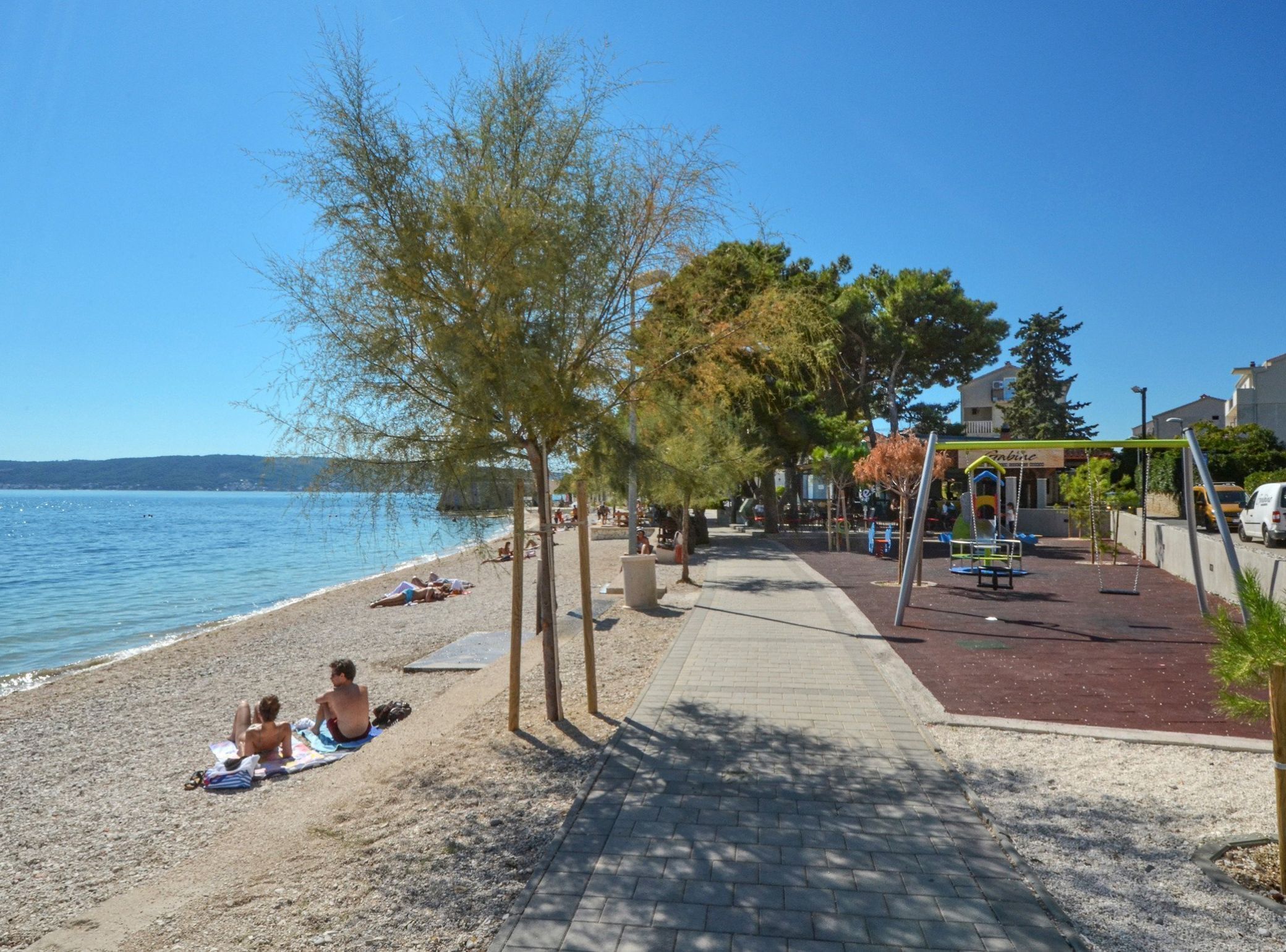 Una playa con gente tomando el sol, un parque infantil y un cielo azul despejado.