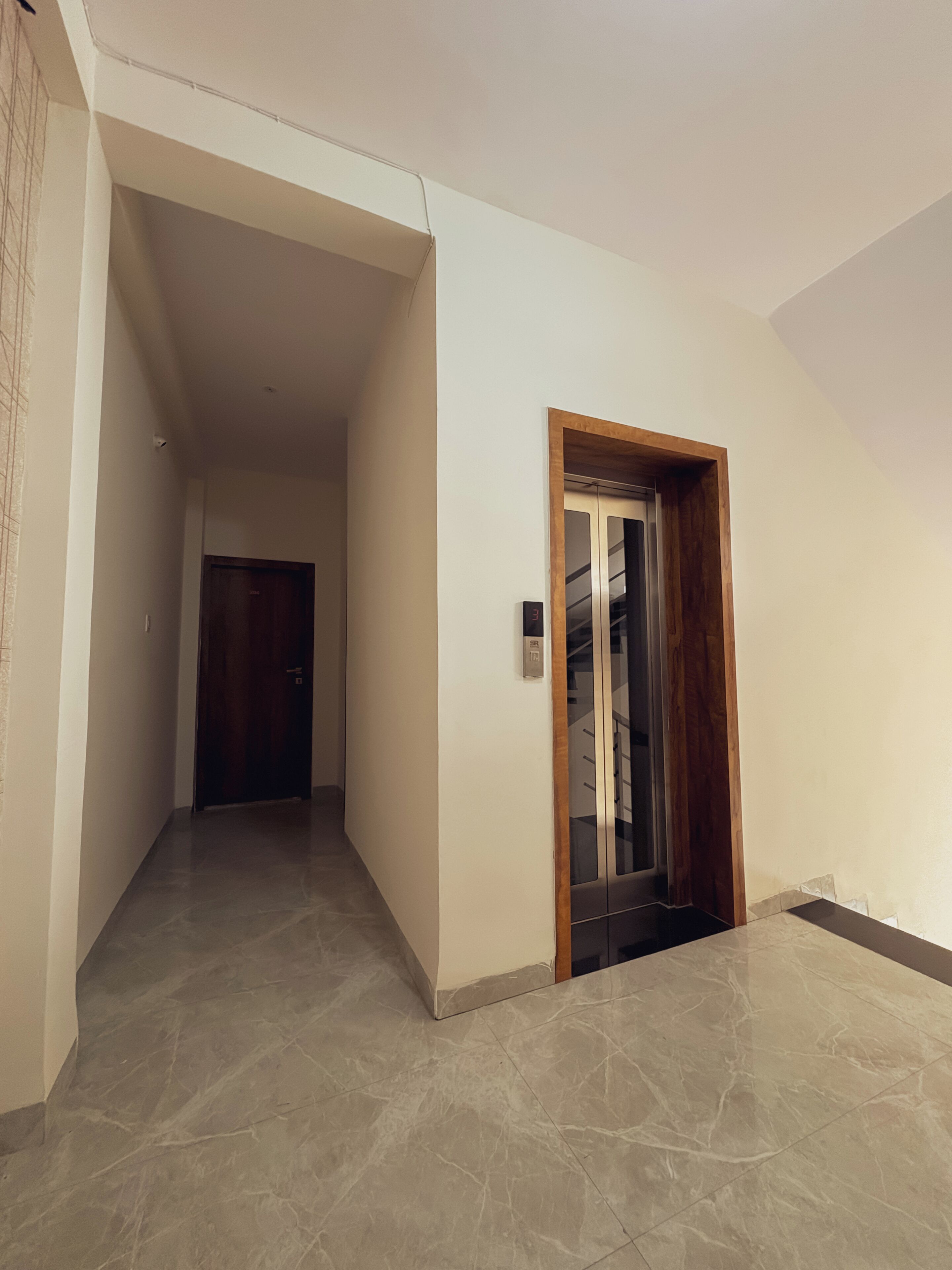 A hallway with a wooden-framed door leading to a room, a tiled floor, and a plain white wall.