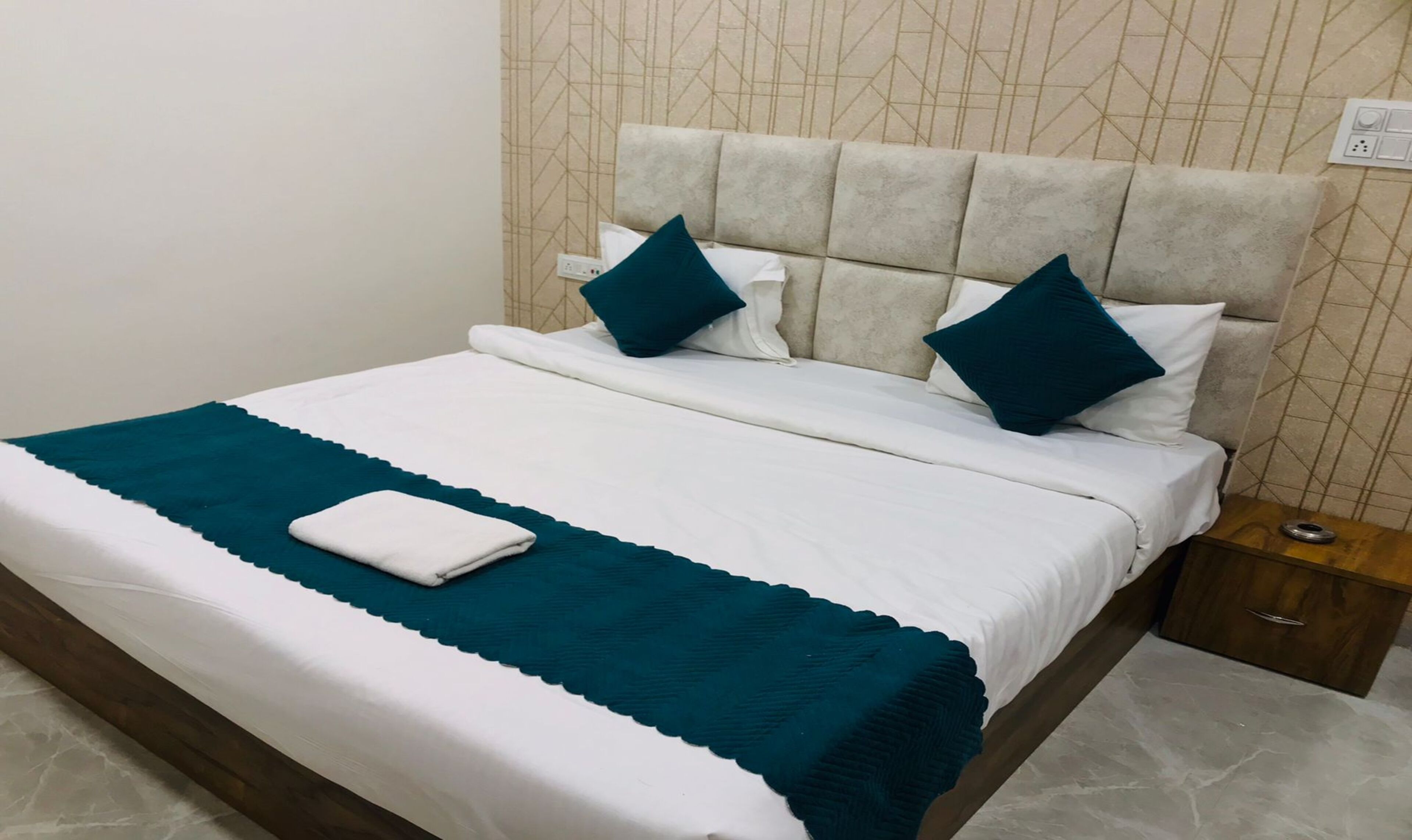 Una cama bien tendida con una colcha blanca y turquesa, dos almohadas turquesas y una toalla blanca sobre la cama.
