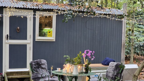 A small outdoor seating area with a table and chairs, surrounded by a dark wooden structure with a window and a door. - If you go into the woods today... Off-grid yet comfy, warm and luxurious  (Canterbury)