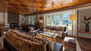 A spacious living room with a wooden ceiling, large windows, a glass coffee table, and a fireplace. - Chalet Les Ecureuils (Zermatt)