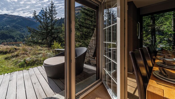 A modern outdoor dining area with a wooden deck, a glass door leading to a scenic mountain view, and a set table with chairs. - Chalet Les Ecureuils (Zermatt)