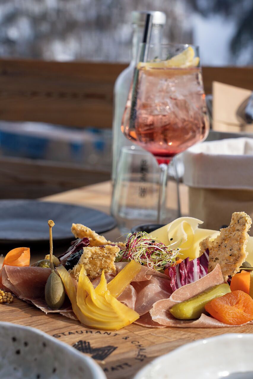 Una tabla de fiambres con diversos embutidos, quesos y verduras, acompañada de una copa de vino rosado.