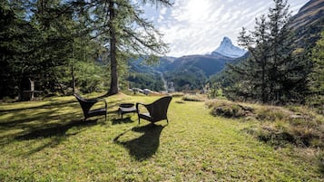 A grassy outdoor area with two black chairs and a small table, surrounded by trees and a mountain view.