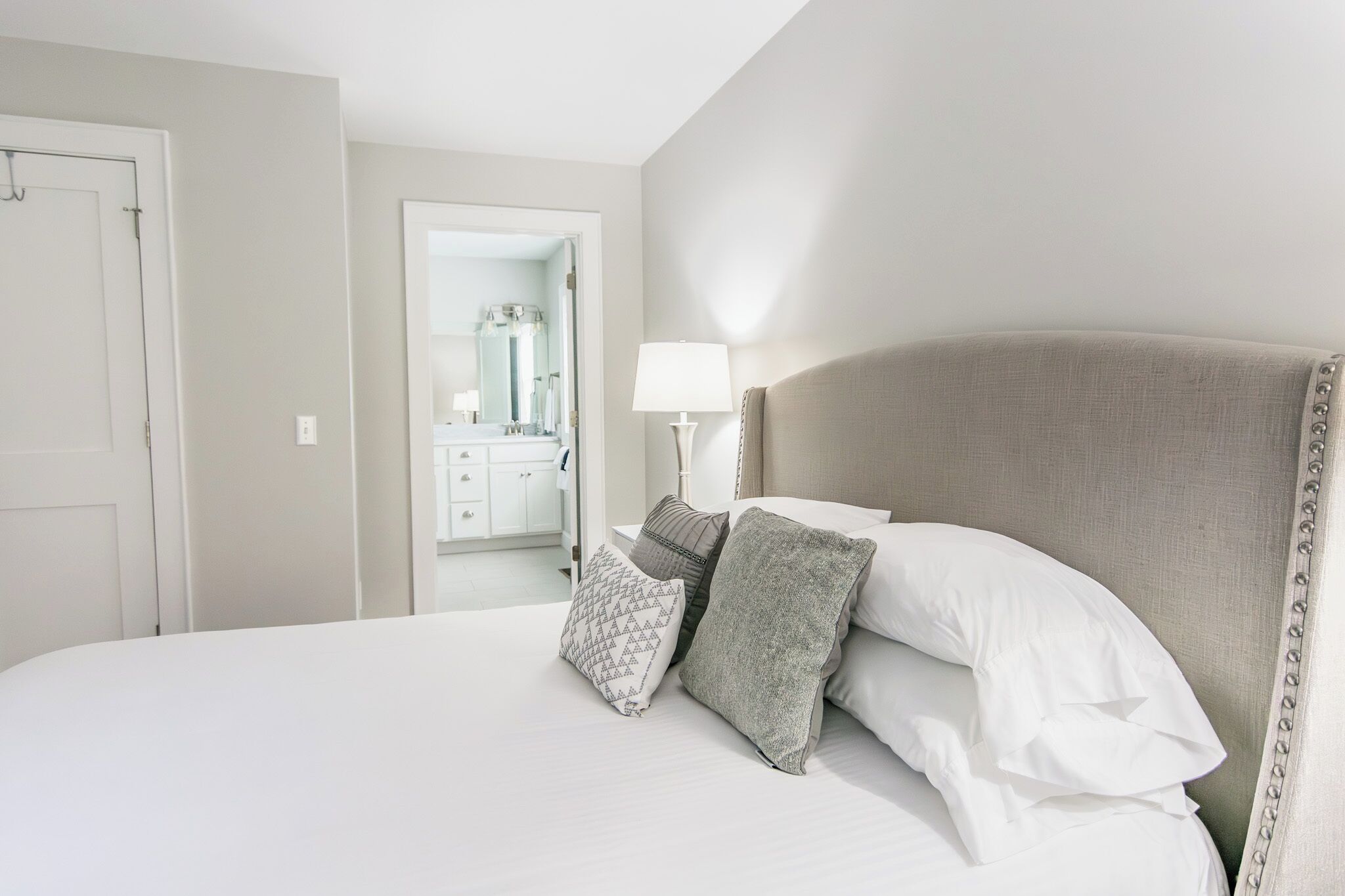 A neatly made bed with white and grey pillows, a bedside lamp, and a view into a bathroom with a mirror and a sink.