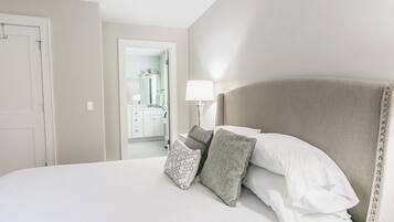 A neatly made bed with white and grey pillows, a bedside lamp, and a view into a bathroom with a mirror and a sink.
