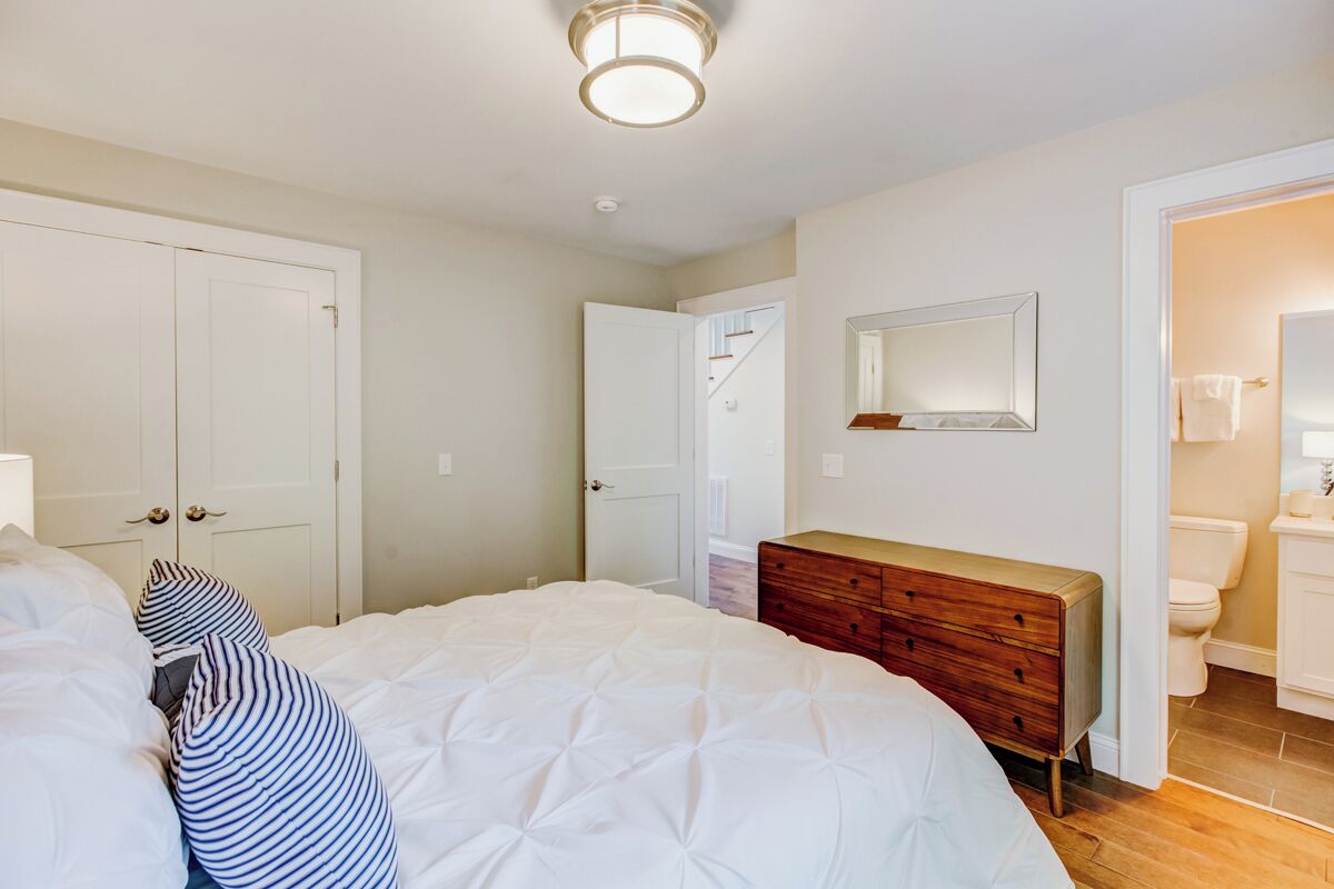 Une chambre à coucher avec un lit, une commode en bois, une porte blanche et une salle de bain visible à travers une porte ouverte.