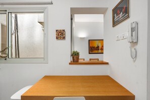 A room with a wooden table, a potted plant, a framed picture, a wall-mounted phone, and a window with blinds.