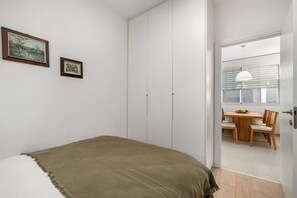 A bedroom with a bed, a wardrobe, a dining area with a table and chairs, and a window with blinds.