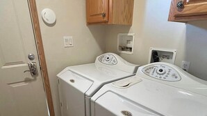 A compact laundry room with two white front-loading washers and dryers, wooden cabinets above, and a door with a silver handle. - Spacious Peaceful 2-bedroom home in Saint Ignatius, Montana with amazing views. (Saint Ignatius)