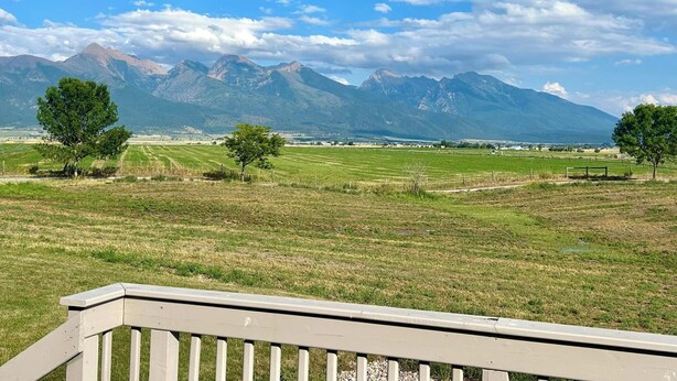 A scenic view from a balcony with a wooden railing, overlooking a grassy field and distant mountains under a partly cloudy sky. - Spacious Peaceful 2-bedroom home in Saint Ignatius, Montana with amazing views. (Saint Ignatius)
