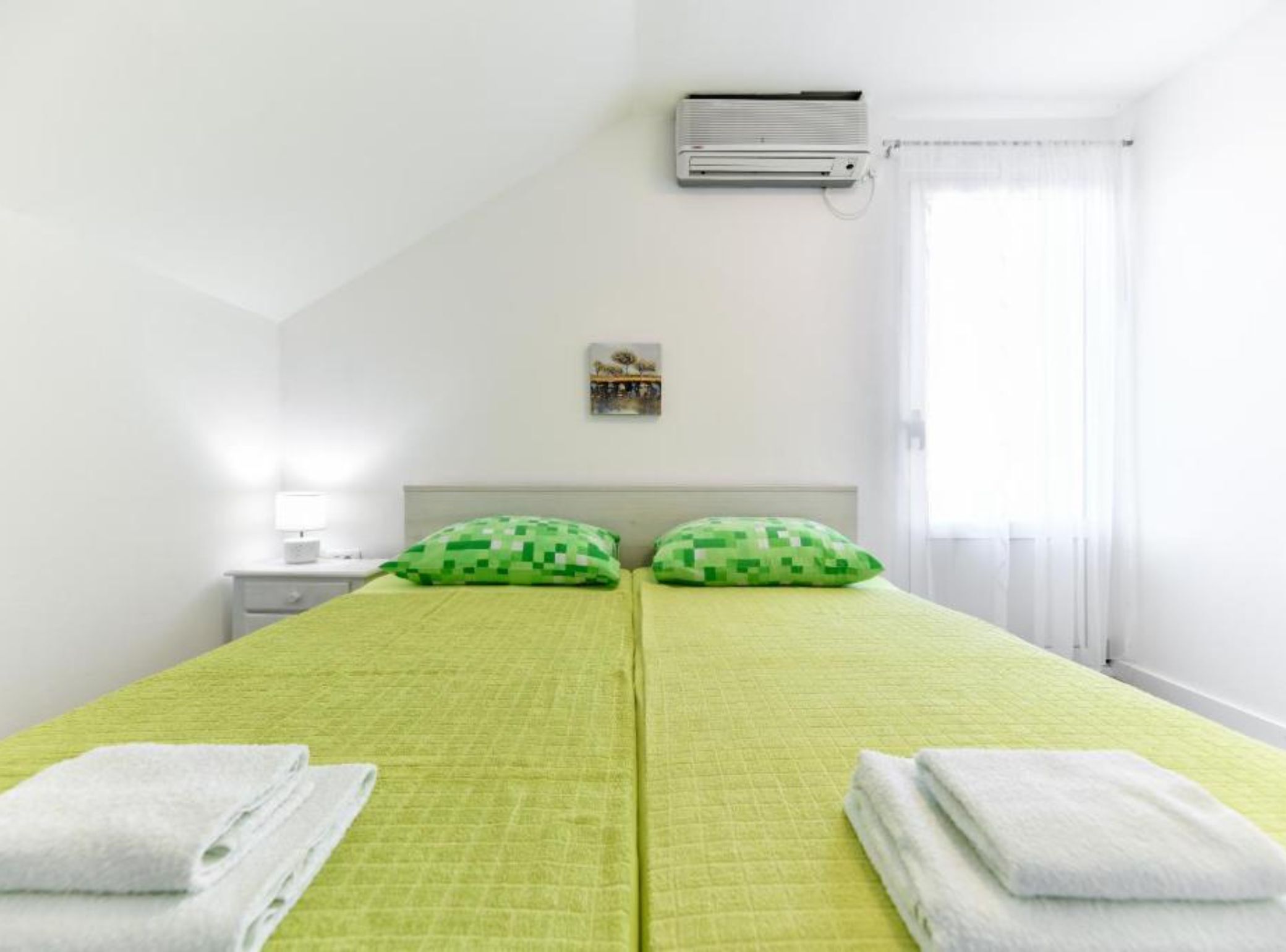 A room with a bed, two pillows, a nightstand, a lamp, an air conditioner, and a window with curtains.