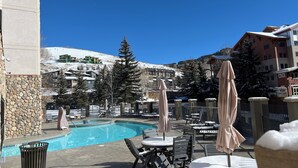 A pool area with lounge chairs and umbrellas, surrounded by buildings and snow-covered hills.