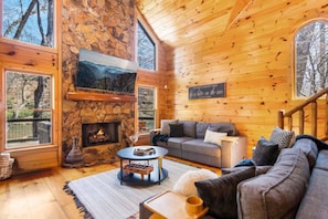 A cozy cabin interior with a stone fireplace, wooden walls, a sofa, and large windows offering views of the outdoors. - Riverfront Cabin in Coosawatte with Playset (Ellijay)
