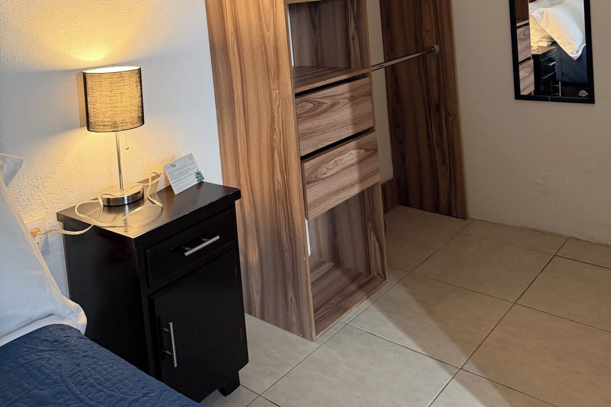 A bedroom with a wooden wardrobe, a bedside table with a lamp, a mirror, and a bed.