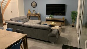 Een woonkamer met een grijze bank, een houten salontafel, een aan de muur bevestigde televisie en een trap naar een andere kamer.