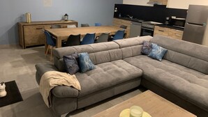 Een moderne woonkamer met een grijze hoekbank, een houten eettafel en een keuken met een zwart aanrecht en roestvrijstalen apparatuur.