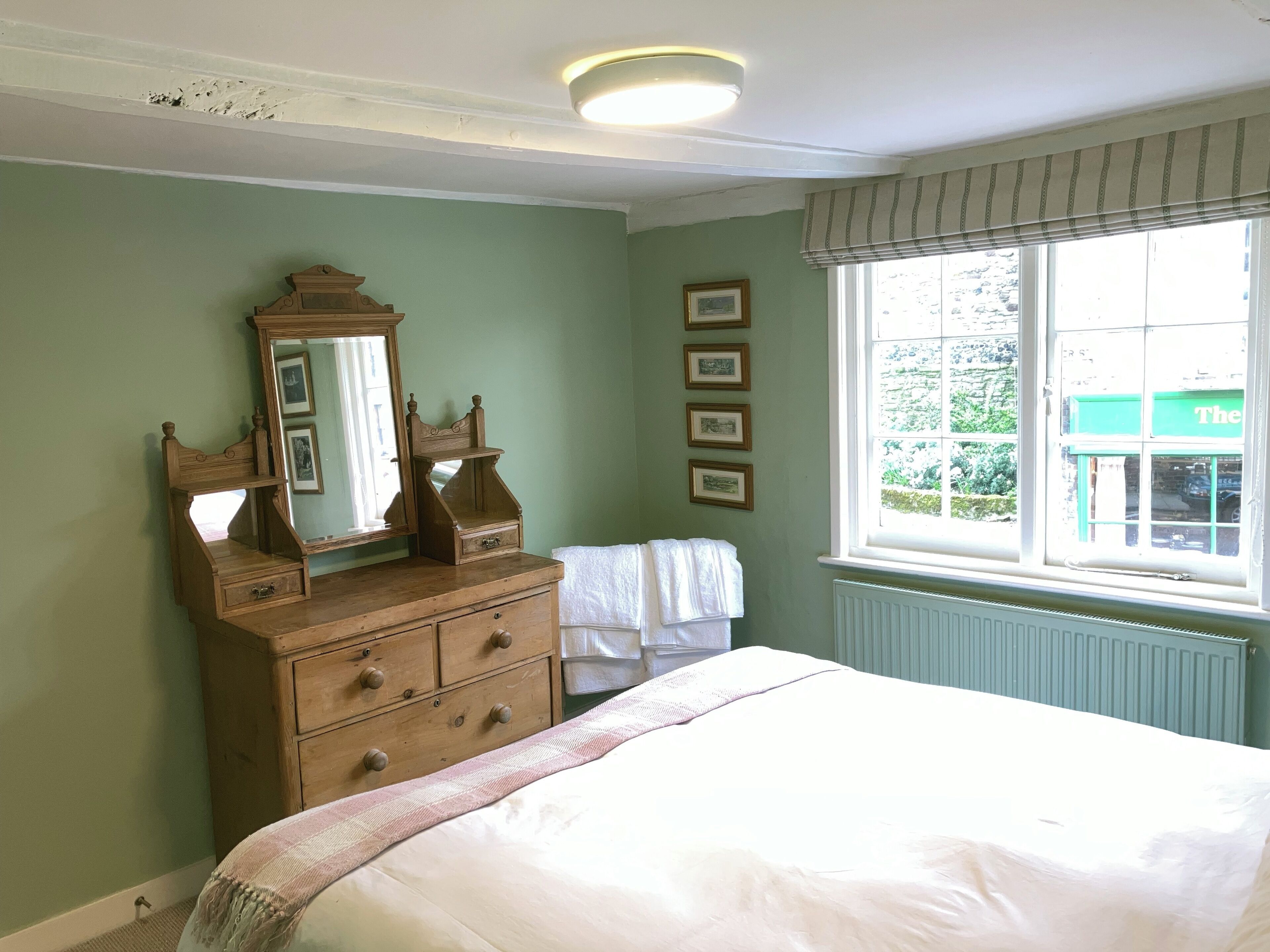 Une chambre à coucher comprenant un lit, une commode, un miroir, des tableaux encadrés et une fenêtre donnant sur un bâtiment verdoyant.