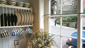Een keuken met witte kastjes, een raam met uitzicht op een stenen muur en een straat, en een vaas met bloemen op het aanrecht.