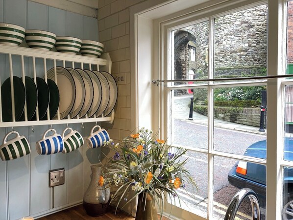 A kitchen with white cabinets, a window with a view of a stone wall and a street, and a vase with flowers on the counter. - Landgate Cottage is opposite the 14th Century entrance to historic Rye town (Rye)