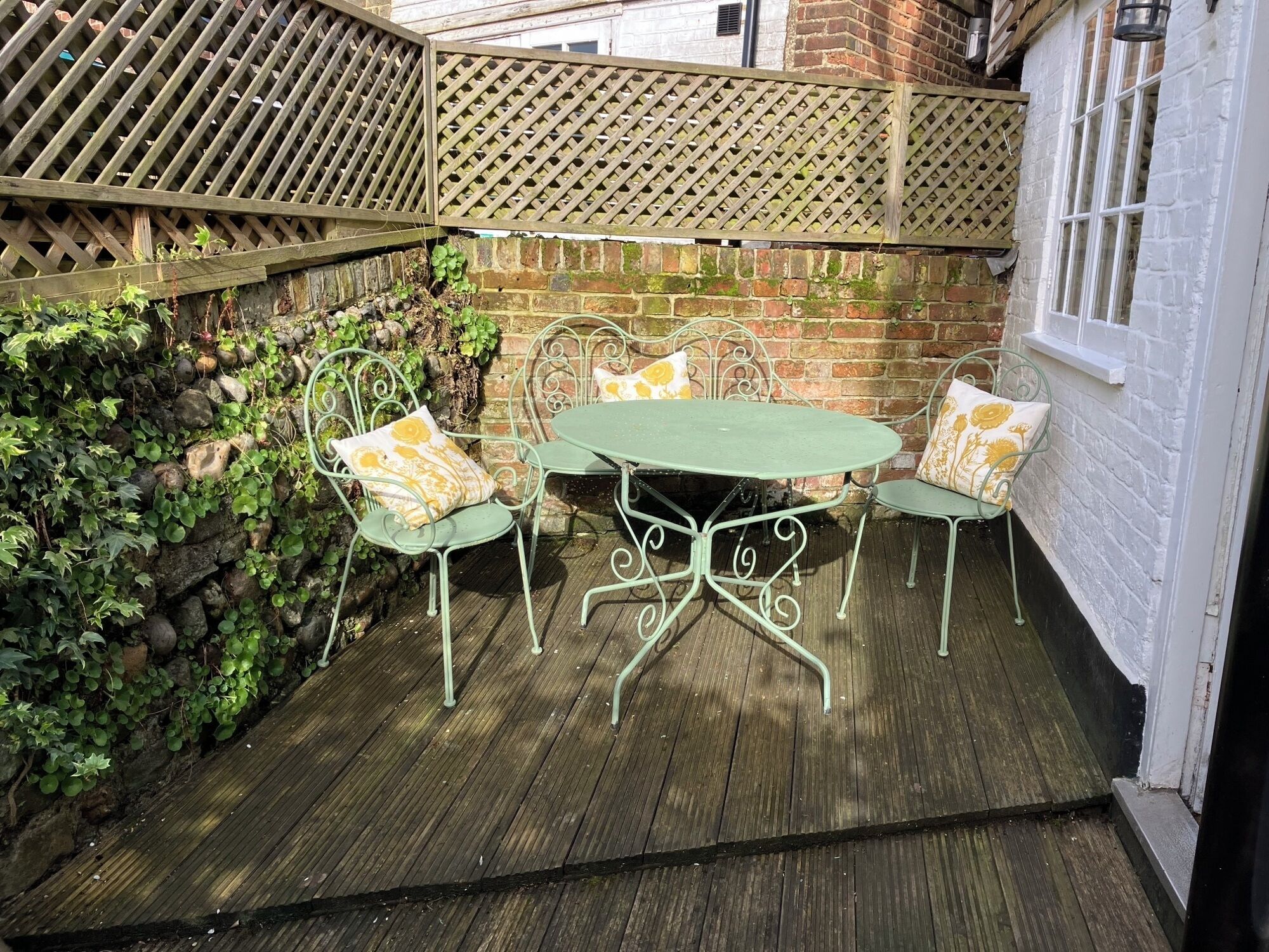 Een terras met een groene metalen tafel en stoelen, omgeven door een houten schutting en een bakstenen muur.