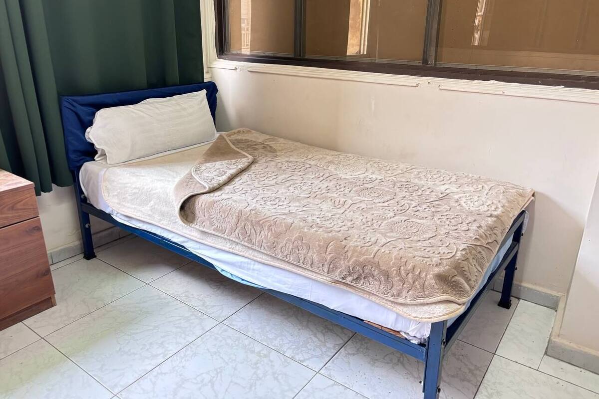 A single bed with a patterned bedspread in a room with a window, a wooden dresser, and tiled flooring.