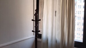 Una habitación con una ventana cubierta por cortinas traslúcidas, un perchero de madera y una silla de mimbre.